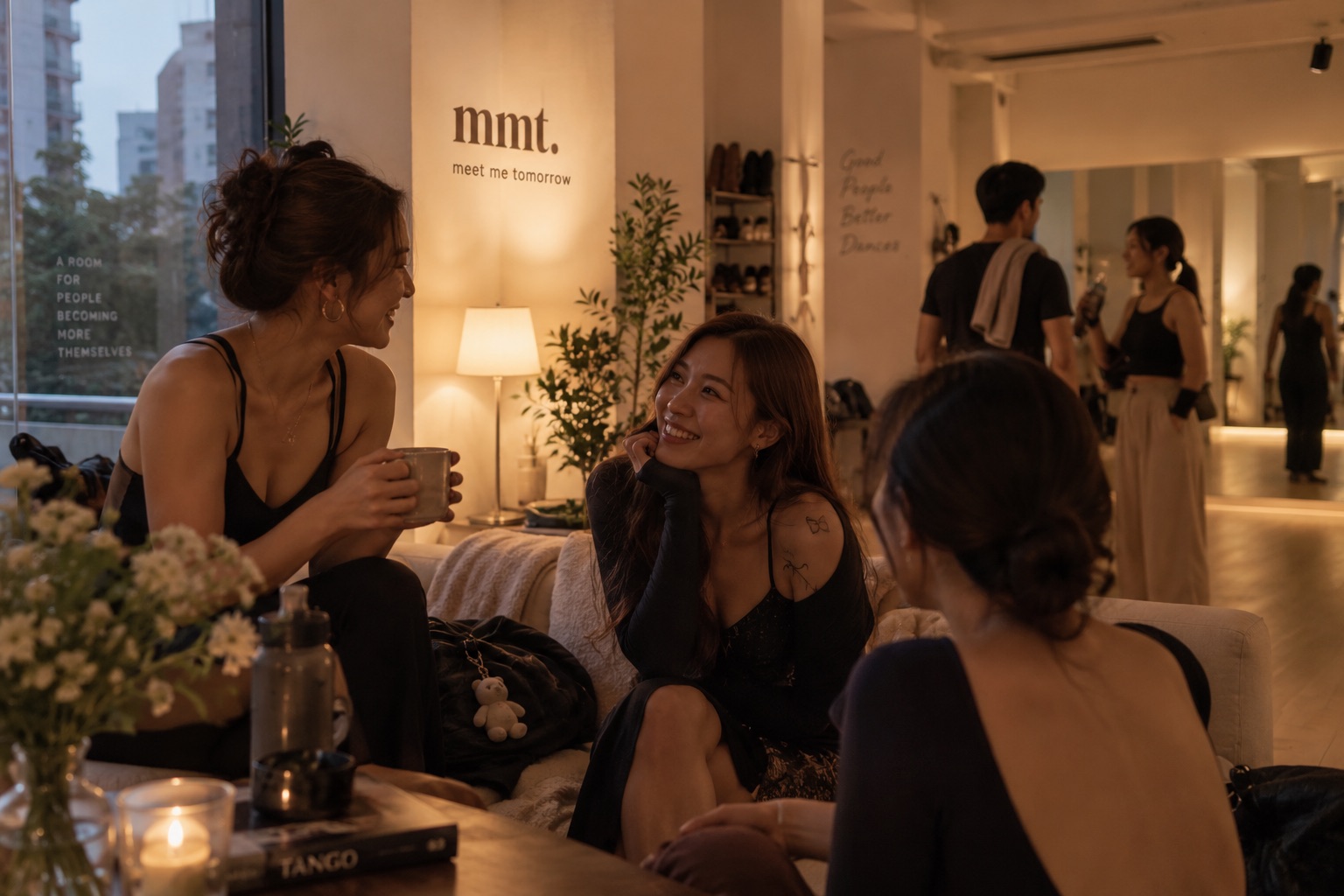 Three people talking on the couch at Meet Me Tomorrow, warm lamplight, others gathered by the mirror behind them